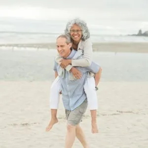 older couple walking on the beach and the man is carrying the woman piggy back